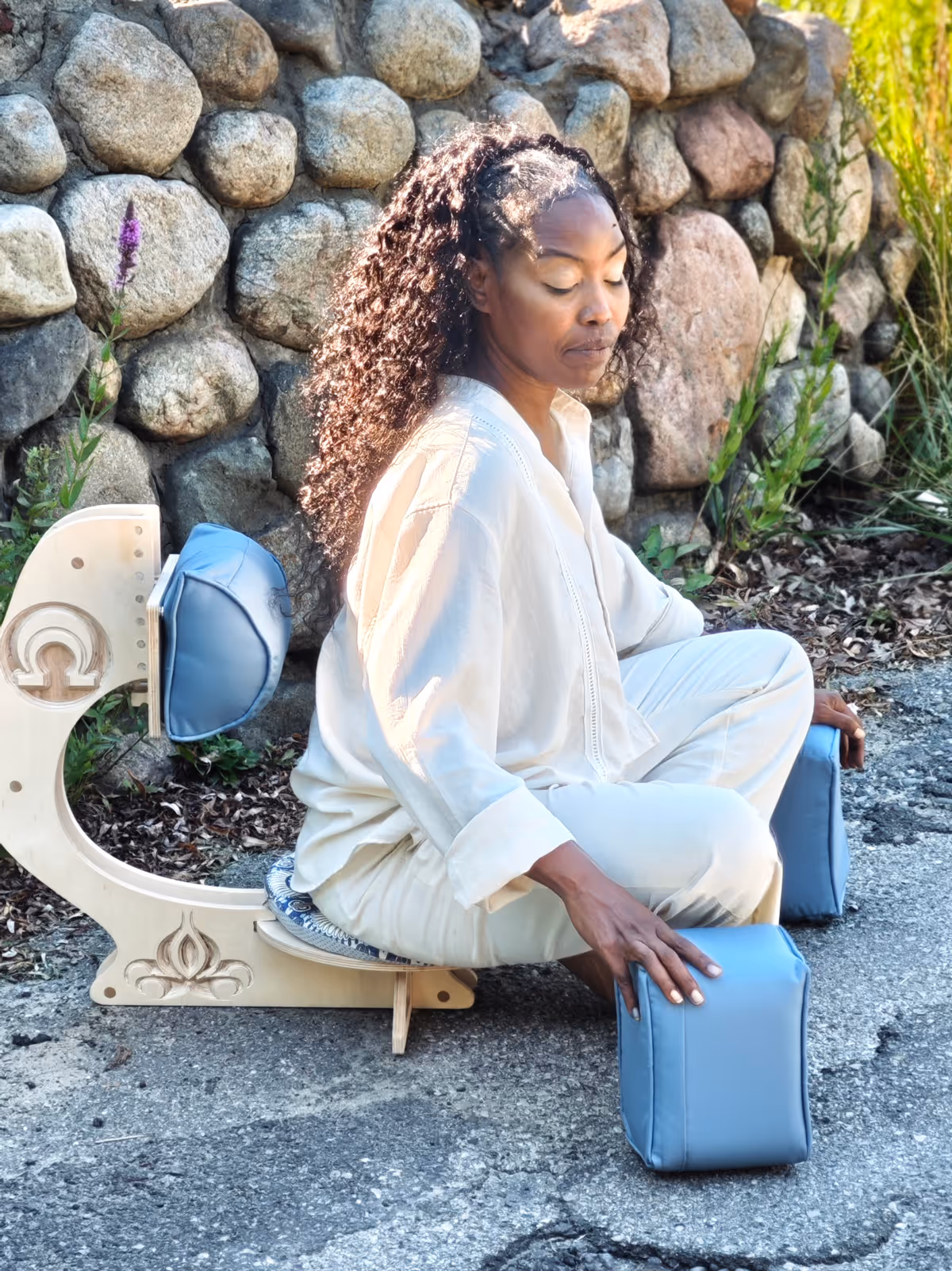 Woman seated in the OM Chair outdoors on a stone patio in a meditative posture, wearing a white outfit, with blue cushion support pads