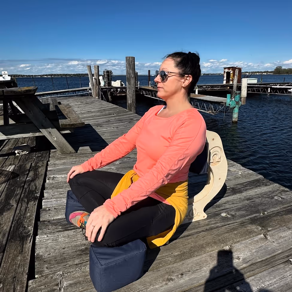 Jennifer, Registered Nurse and Veteran, seated outdoors on a wooden deck using the OM Chair with an orange cushion, wearing a coral jacket