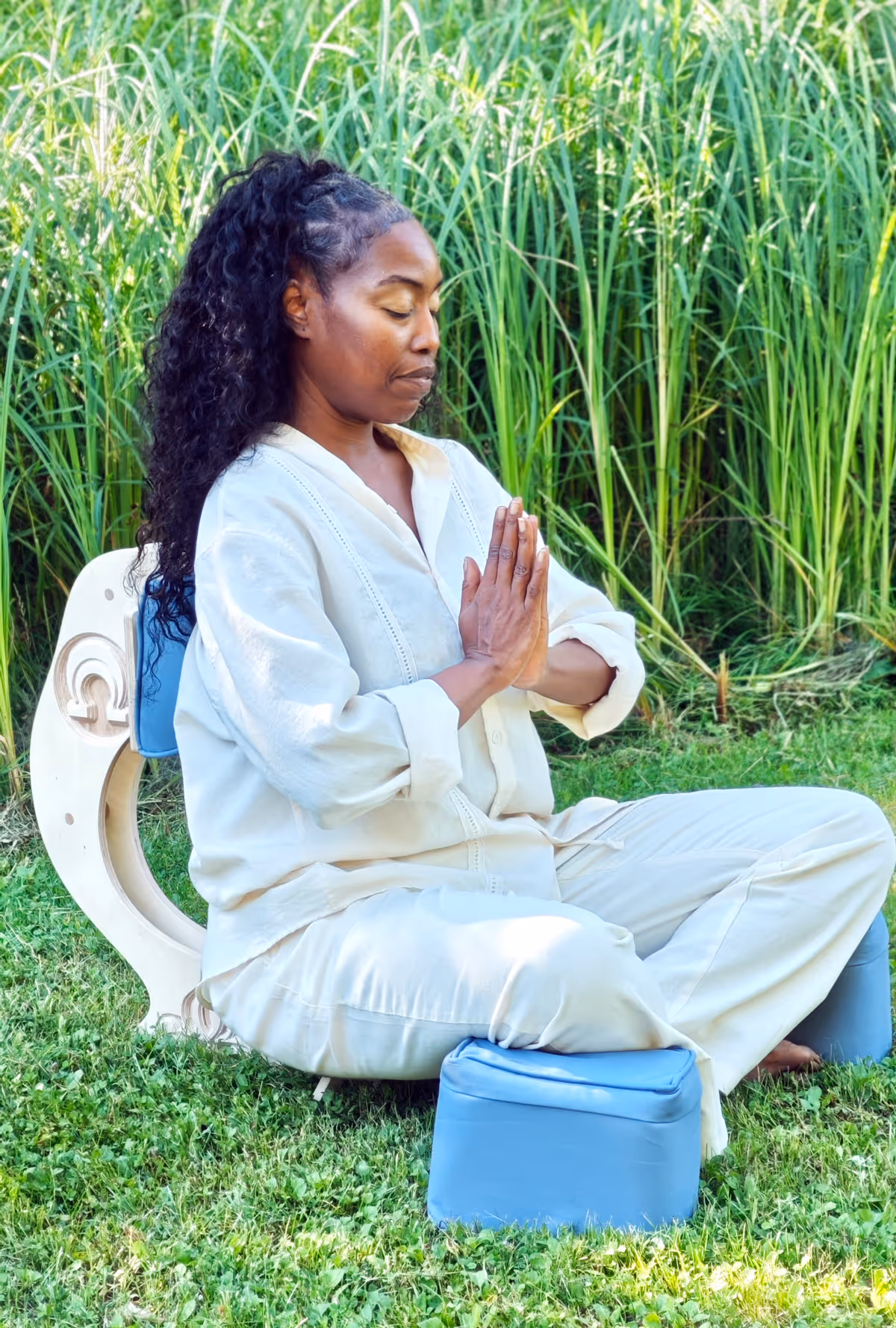 Woman meditating in the OM Chair seated cross-legged in a grassy garden setting, hands pressed together in prayer position